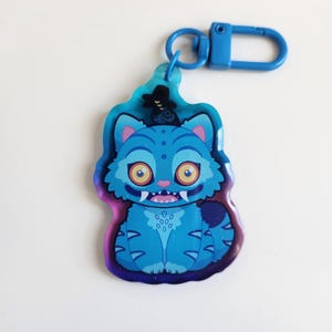 K&ouml;nnte beinhalten: Ein blauer, katzenf&ouml;rmiger Schl&uuml;sselanh&auml;nger mit Cartoon-Design. Die Katze hat gro&szlig;e gelbe Augen, spitze Z&auml;hne und einen kleinen schwarzen Hut. Der Schl&uuml;sselanh&auml;nger hat einen blauen Verschluss.