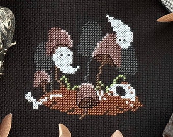 May include: Cross-stitch pattern featuring three brown mushrooms with white ghost faces on a black background. The mushrooms are surrounded by green leaves and white bones.