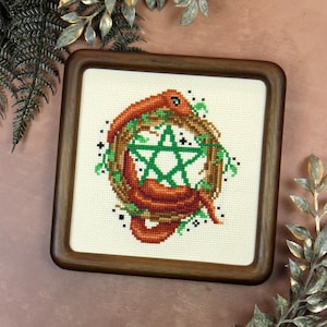 Peut inclure: Broderie au point de croix dans un cadre en bois carré. Le motif représente un serpent brun enroulé autour d'un pentagramme vert, entouré d'une couronne de feuilles vertes et de vignes brunes. Des étoiles noires ornent le design.