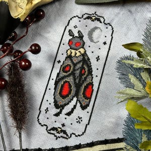 Puede incluir: Patrón de punto de cruz con una polilla negra y roja con una luna creciente y estrellas en el fondo. La polilla tiene un cuello blanco y ojos rojos.