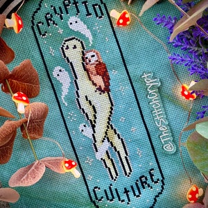 May include: Cross-stitch pattern featuring a green cryptid with a small brown owl on its back. The pattern is on a teal background with white stars and the words "Cryptid Culture" in black. The pattern is surrounded by a black border.