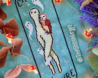Op de afbeelding: Borduurpatroon met een groene cryptide met een kleine bruine uil op zijn rug. Het patroon is op een teal achtergrond met witte sterren en de woorden "Cryptid Culture" in zwart. Het patroon is omgeven door een zwarte rand.