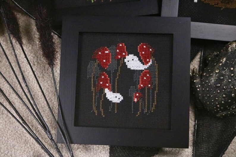 May include: A framed cross-stitch embroidery featuring a dark background with white ghosts and red mushrooms.