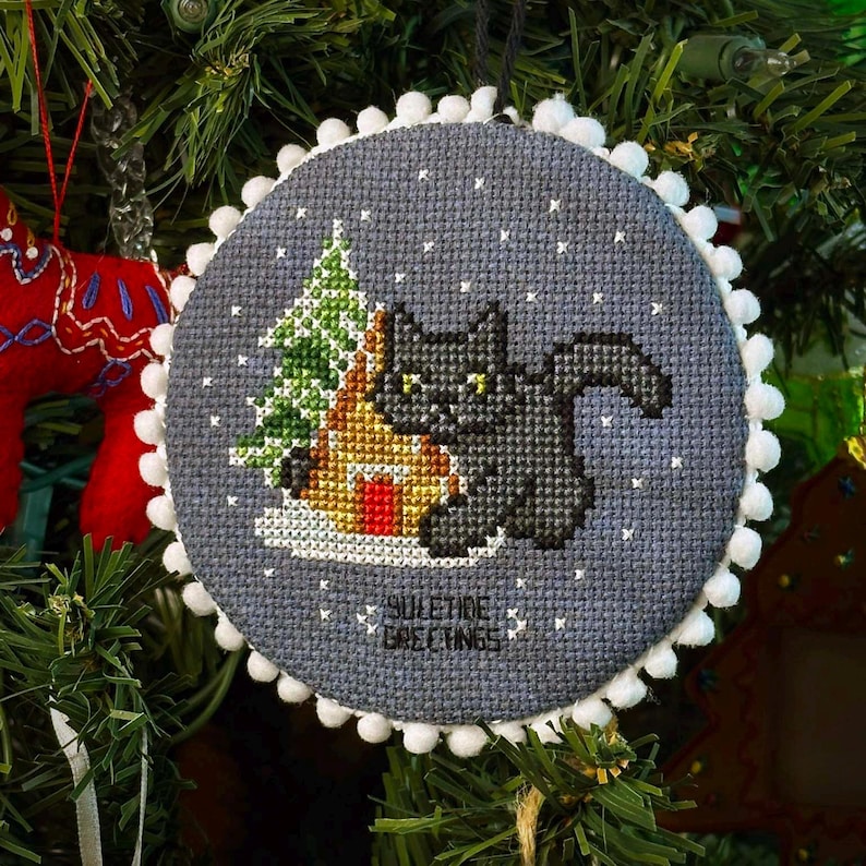 May include: A round, gray felt ornament with a white pom-pom trim. The ornament features a black cat sitting in front of a small, yellow house with a green tree behind it. The text "Suletire Greetings" is embroidered on the ornament.