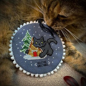 May include: A gray and white round ornament with a black cat sitting in front of a small house and a green Christmas tree. The ornament has white pom-poms around the edge and the text "YULETIDE GREETINGS" is printed on the bottom. A brown tabby cat is lying on a brown surface with its head resting on the ornament.