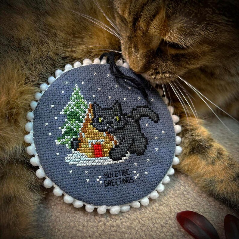 May include: A gray and white round ornament with a black cat sitting in front of a small house and a green Christmas tree. The ornament has white pom-poms around the edge and the text "YULETIDE GREETINGS" is printed on the bottom. A brown tabby cat is lying on a brown surface with its head resting on the ornament.