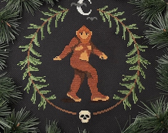 May include: A cross-stitch embroidery design featuring a brown Bigfoot figure walking on a black background with a crescent moon and bats above. The design is surrounded by green pine branches and a skull at the bottom.