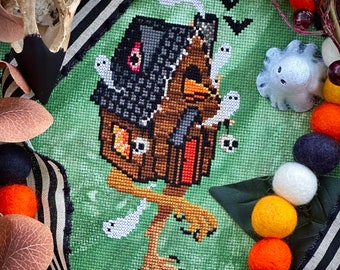 May include: A cross-stitch pattern of a brown house with a black roof and a witch's leg sticking out of the bottom. The house is surrounded by white ghosts and bats.