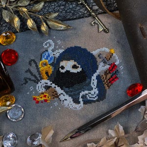 Puede incluir: Diseño de punto de cruz que presenta un ninja con una máscara negra y una capucha azul, rodeado de cadenas, llaves y otros objetos pequeños. El diseño está sobre una tela gris, con elementos decorativos como gemas y un bolígrafo.