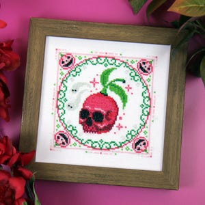 May include: Cross-stitch art in a wooden frame. The design features a pink skull with green leaves, surrounded by a green and pink border. Two white ghost-like figures are on the left side of the skull. The background is white.