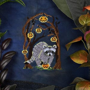 Op de afbeelding: Kruissteekontwerp met een wasbeer die een pompoen vasthoudt, in een Halloween-scène. Het kunstwerk toont een boom met hangende pompoenlantaarns en vleermuizen, allemaal in grijstinten, bruin en oranje tegen een donkerblauwe achtergrond.