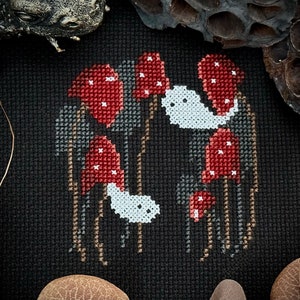 May include: Cross-stitch embroidery of a spooky forest scene with red mushrooms and white ghosts. The mushrooms have white spots and the ghosts have black eyes. The background is black.