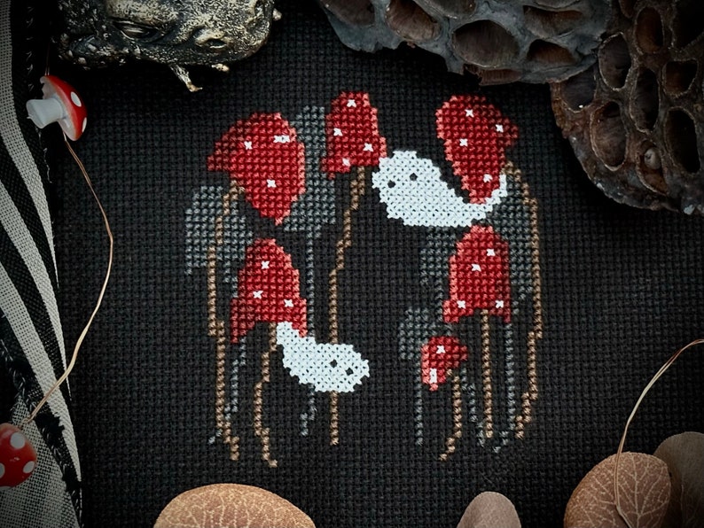 May include: Cross-stitch embroidery of a spooky forest scene with red mushrooms and white ghosts. The mushrooms have white spots and the ghosts have black eyes. The background is black.