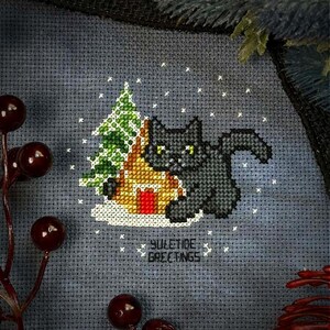 May include: Cross-stitch pattern featuring a black cat sitting next to a gingerbread house with a green Christmas tree in the background. The design is on a gray fabric with white snowflakes and the text "YULETIDE GREETINGS".