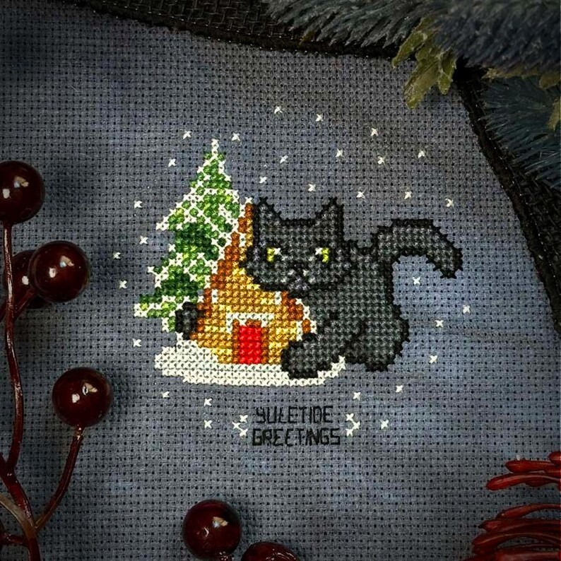 May include: Cross-stitch pattern featuring a black cat sitting next to a gingerbread house with a green Christmas tree in the background. The design is on a gray fabric with white snowflakes and the text "YULETIDE GREETINGS".