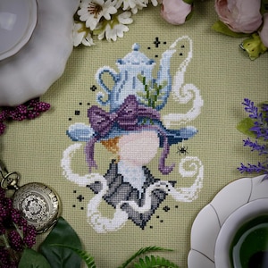 May include: Cross-stitch design featuring a whimsical figure with a teapot hat, adorned with a purple bow and swirling white accents. The design includes a small spider and is set against a light green background, surrounded by floral elements.