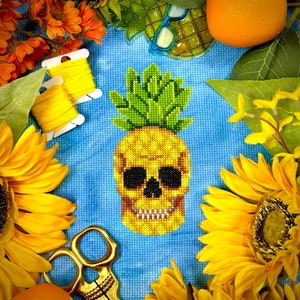 May include: A cross-stitch pattern of a pineapple with a skull face on a blue background. The pineapple is yellow and green, and the skull is white with black eyes. The background is a light blue.