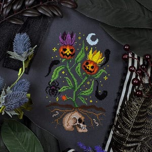 May include: Cross-stitch design depicting a Halloween plant with jack-o'-lantern flowers, a skull, and a spider. The design includes a crescent moon and other spooky elements, all stitched on a dark grey fabric.
