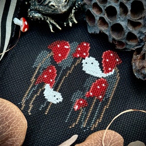 May include: Cross-stitch embroidery of red mushrooms with white spots on a black fabric background. The mushrooms are arranged in a cluster with some taller than others. The embroidery is surrounded by brown leaves and a dried lotus pod.