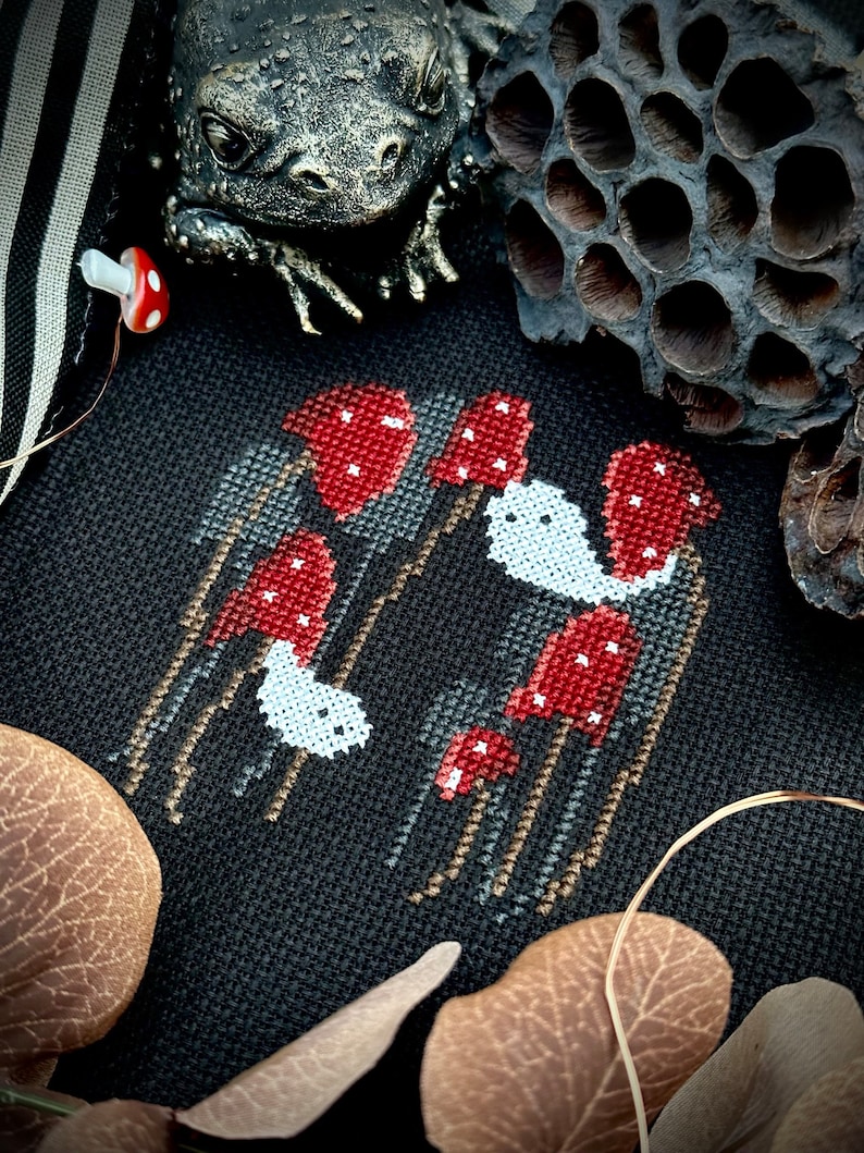 May include: Cross-stitch embroidery of red mushrooms with white spots on a black fabric background. The mushrooms are arranged in a cluster with some taller than others. The embroidery is surrounded by brown leaves and a dried lotus pod.