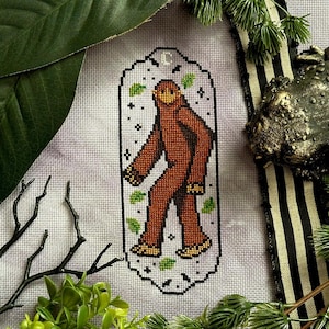 Op de afbeelding: Borduurpatroon van een bruine Bigfoot die door een bos loopt. Het patroon is ingelijst in een witte ovaal met een zwarte omtrek. De achtergrond is lichtpaars.