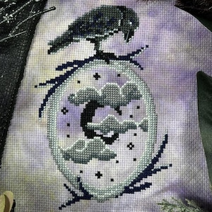 May include: A cross-stitch embroidery design featuring a black raven perched on a branch, with a crescent moon and clouds in a circular frame. The background is a light purple with a subtle watercolor effect.
