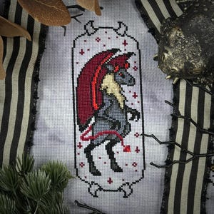 May include: Cross-stitch pattern of a gray and red demon with wings and a tail, framed in a white bone-shaped border. The background is a light gray with white stars.