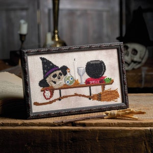 Puede incluir: Un diseño de punto de cruz enmarcado que presenta un caldero de bruja, una calavera con un sombrero de bruja, un libro titulado "Spells" y una escoba.