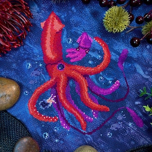 May include: A cross-stitch embroidery of a red squid with a smaller pink squid in its tentacles. The squid are surrounded by blue and white bubbles on a dark blue background.