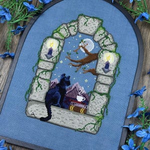 Puede incluir: Arte de punto de cruz que presenta un gato negro, una luna llena y un árbol dentro de un arco de piedra. La escena incluye velas encendidas y un pequeño cofre. El fondo es de tela azul claro y la obra está enmarcada.