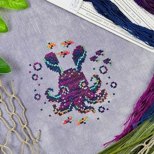 Puede incluir: Punto de cruz de un pulpo morado con detalles turquesa y naranja, rodeado de burbujas y peces pequeños. El pulpo está sobre una tela morada clara, con madejas de hilo azul y morado.