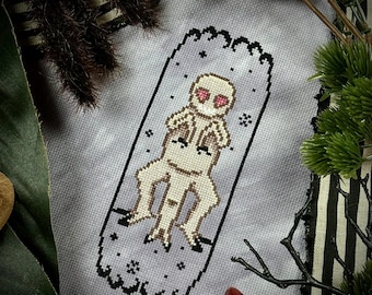 May include: Cross-stitch pattern of a pale-skinned creature with large eyes and a toothy grin, sitting on a throne. The creature is surrounded by a starry sky.