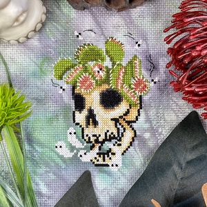 May include: Cross-stitch design featuring a skull with a Venus flytrap crown and small ghost figures. The skull is beige with black eyes and nose. The flytrap is green and red. Small black flies are also stitched into the design.