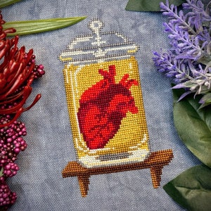 May include: A cross-stitch design of a heart in a glass jar on a wooden stand. The heart is red and the jar is yellow with a white lid.