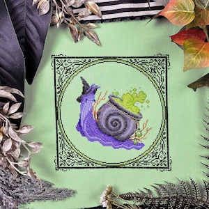 May include: Cross-stitch design featuring a purple snail wearing a witch hat, with a cauldron of green bubbling potion. The design is framed in a black ornate border on a light green background. Decorative leaves surround the cross-stitch.