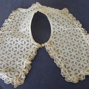 May include: A cream-coloured lace collar with a floral openwork pattern and ruffled edges. The vintage collar is displayed on a dark blue surface, highlighting its delicate design. A classic accessory, perfect for adding a touch of elegance.