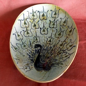 May include: Oval ceramic plate with a peacock design. The peacock is depicted with a black body and a fanned tail in shades of blue and green. The plate has a light blue background and a gold rim.
