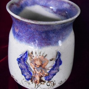 May include: A white ceramic mug with a purple and blue glaze. The mug has a raised design of an angel with wings and a floral pattern.