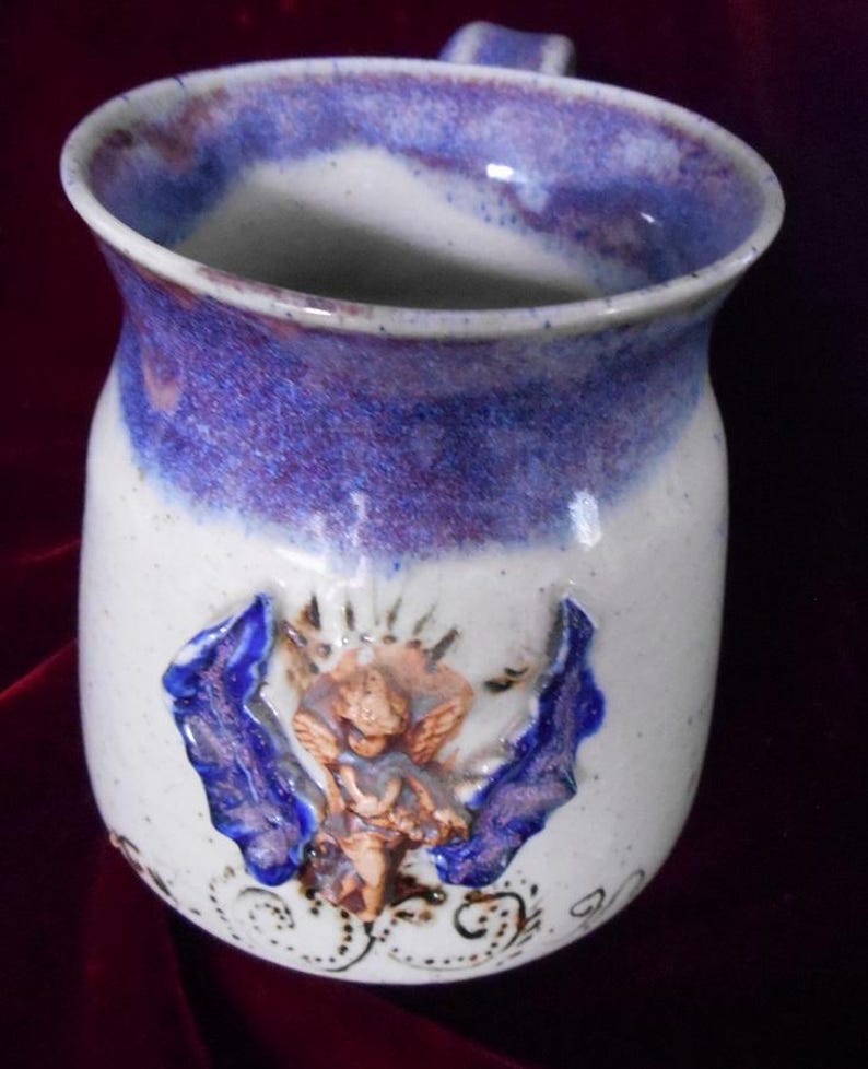 May include: A white ceramic mug with a purple and blue glaze. The mug has a raised design of an angel with wings and a floral pattern.
