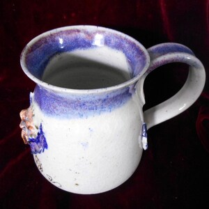May include: White ceramic mug with a blue and purple rim and a floral design. The mug has a handle and is speckled with brown flecks.
