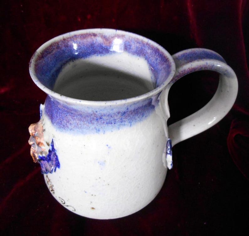 May include: White ceramic mug with a blue and purple rim and a floral design. The mug has a handle and is speckled with brown flecks.