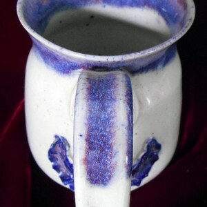 May include: White ceramic mug with a blue and purple glaze. The mug has a handle and a decorative design on the side.