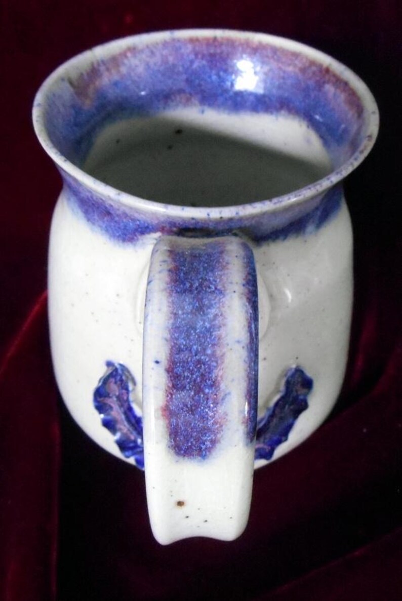 May include: White ceramic mug with a blue and purple glaze. The mug has a handle and a decorative design on the side.
