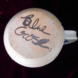 May include: A white ceramic mug with a blue and white design. The bottom of the mug has the signature "Baline Gase" written in black.