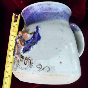 May include: A white ceramic mug with a blue and purple glaze. The mug has a unique handle and a decorative design of a bird with gold accents.