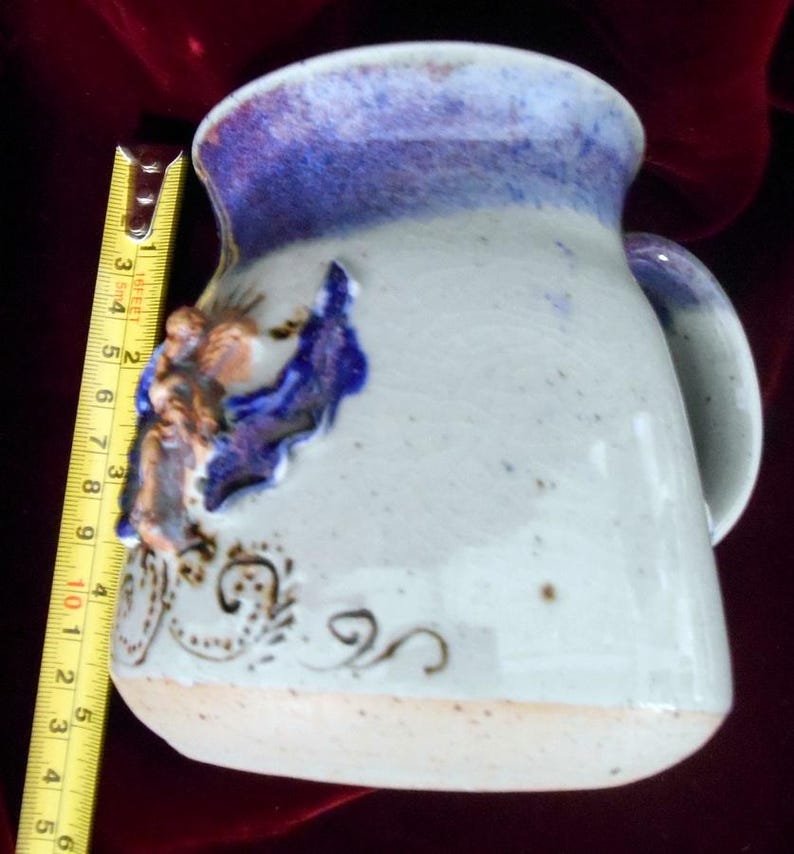 May include: A white ceramic mug with a blue and purple glaze. The mug has a unique handle and a decorative design of a bird with gold accents.