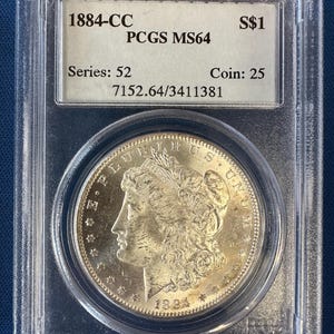 May include: An 1884-CC silver coin, graded PCGS MS64, presented in a protective case. The coin shows a woman's profile and the inscription "E PLURIBUS UNUM". The case also displays "S$1", "Series: 52", and "Coin: 25".