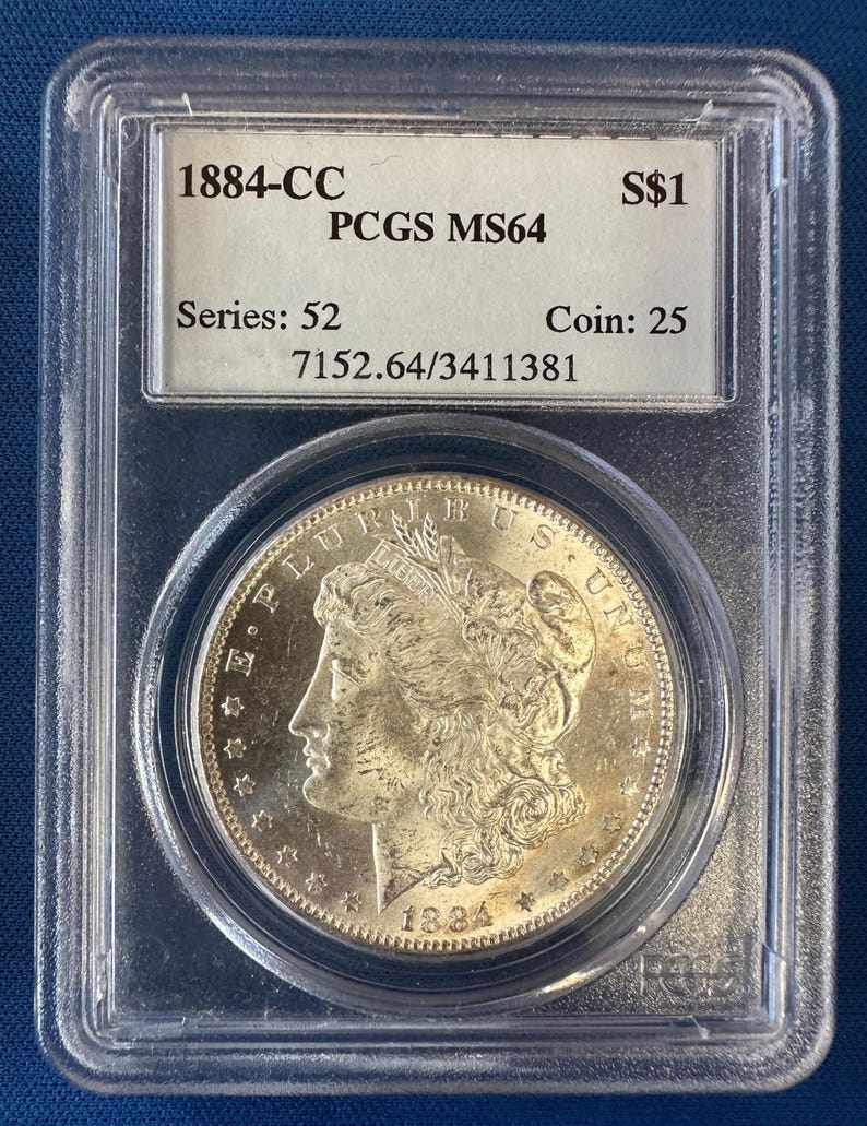 May include: An 1884-CC silver coin, graded PCGS MS64, presented in a protective case. The coin shows a woman's profile and the inscription "E PLURIBUS UNUM". The case also displays "S$1", "Series: 52", and "Coin: 25".