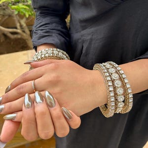 May include: Gold-toned bangles and a bracelet with clear, square and round-cut stones. A small gold ring is also visible. The person is wearing a black long-sleeve shirt. The nails are painted a metallic gold color.