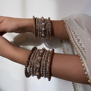 Peut inclure: Un ensemble de bracelets dorés avec des perles roses et blanches. Les bracelets sont empilés les uns sur les autres et sont portés au poignet.
